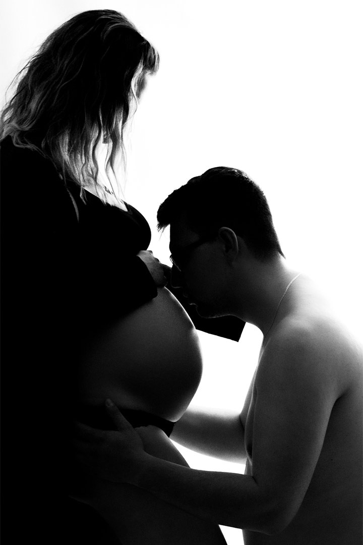 Czarno-białe zdjęcie sylwetki kobiety w ciąży z mężczyzną całującym jej brzuch, oświetlone od tyłu, tworzące silny kontrast.