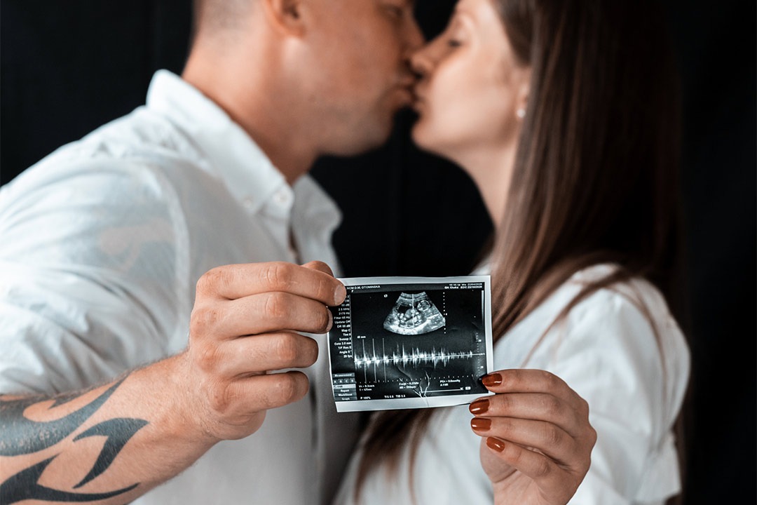 Zakochana para w białych koszulach całuje się, trzymając zdjęcie USG w dłoniach; widoczny tatuaż na ramieniu mężczyzny.