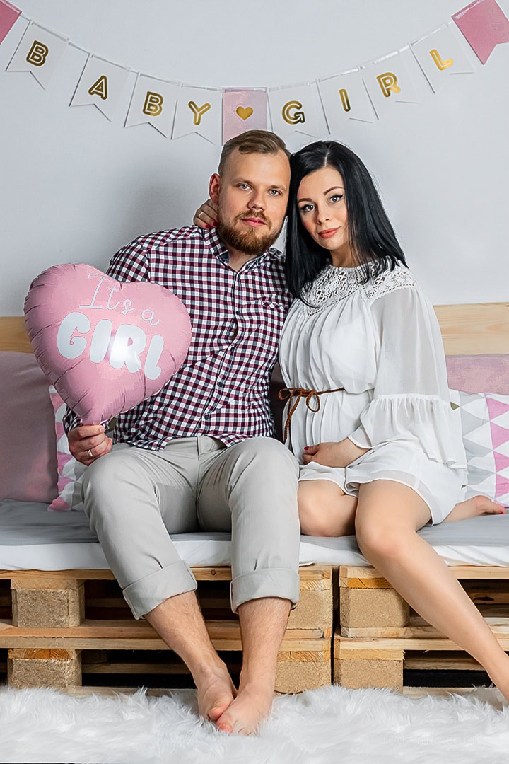 Uśmiechnięta para: przyszła mama w białej sukience z brązowym paskiem i partner trzymający różowy balon w kształcie serca z napisem 'It's a GIRL', siedzą na łóżku z palet pod banerem 'BABY GIRL'.
