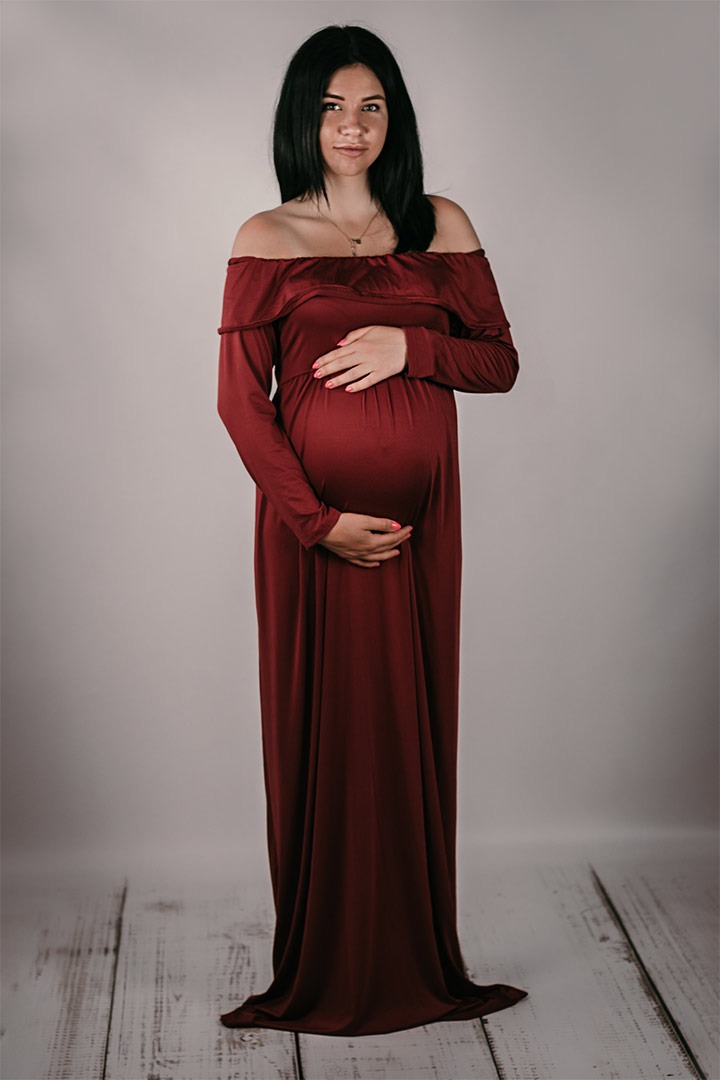 Kobieta w ciąży, ubrana w bordową sukienkę z odkrytymi ramionami, delikatnie trzyma ręce na brzuchu, pozując na tle jasnoszarej ściany i podłogi imitującej bielone drewno.