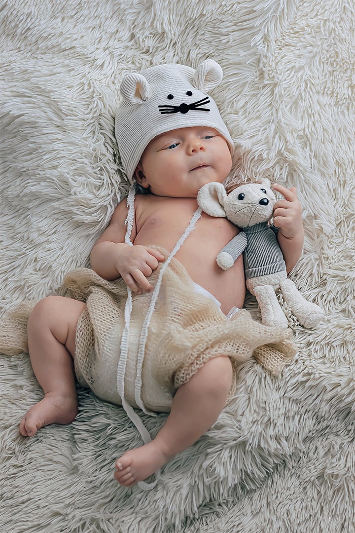 Noworodek leży na puszystym kocu w czapce z uszami myszki, trzymając pluszową myszkę w paski.