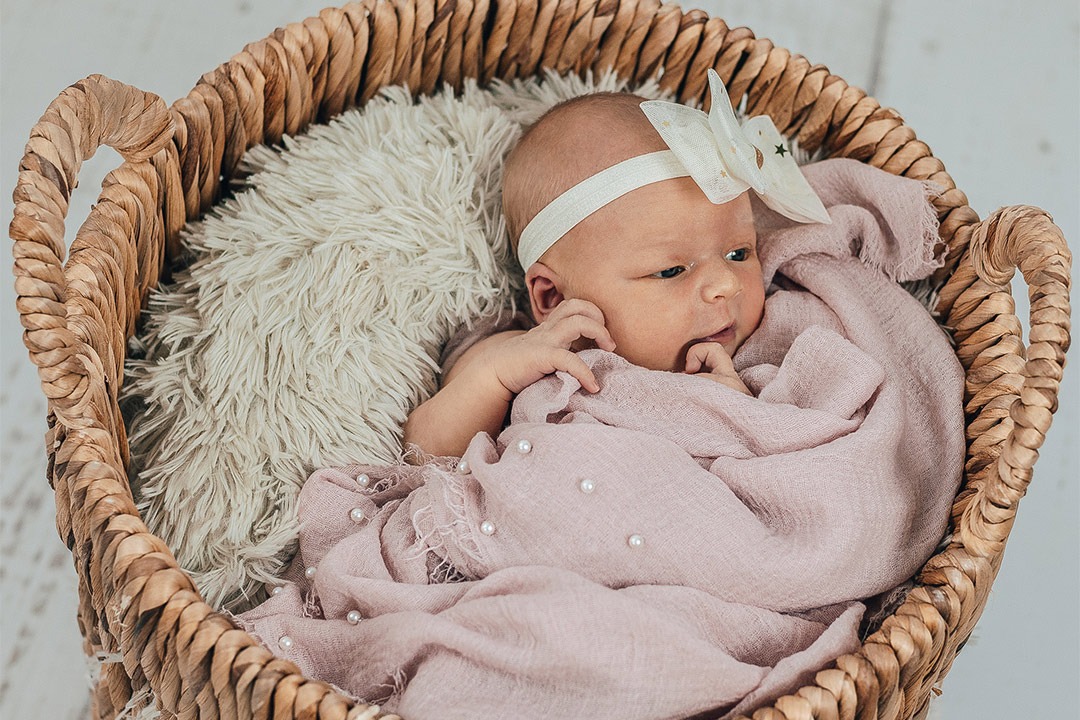 Noworodek w koszu wiklinowym, owinięty w różowy muślin z drobnymi perłami, na białym futrzanym kocyku, z opaską z kokardką w gwiazdki.