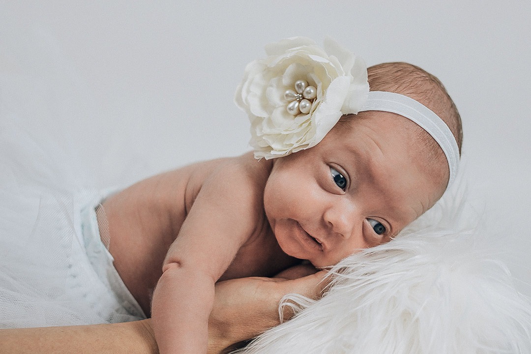 Noworodek w białej opasce z kwiatem, leżący na puszystym, białym futrze, wsparty na dłoni dorosłego.