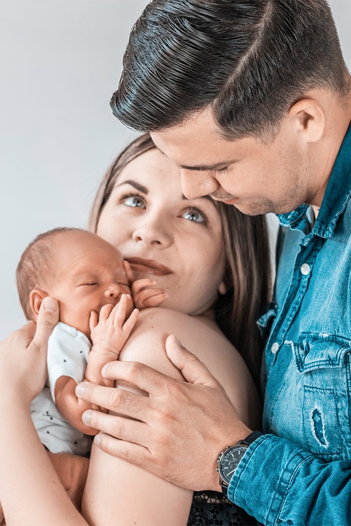 Rodzinne ujęcie: rodzice trzymają noworodka, matka spogląda w górę, ojciec patrzy na dziecko, na nadgarstku ojca widoczny zegarek.