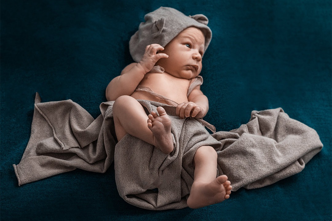 Noworodek leży na ciemnym tle, ubrany w szary strój z uszami, z uniesioną rączką i spojrzeniem w bok.