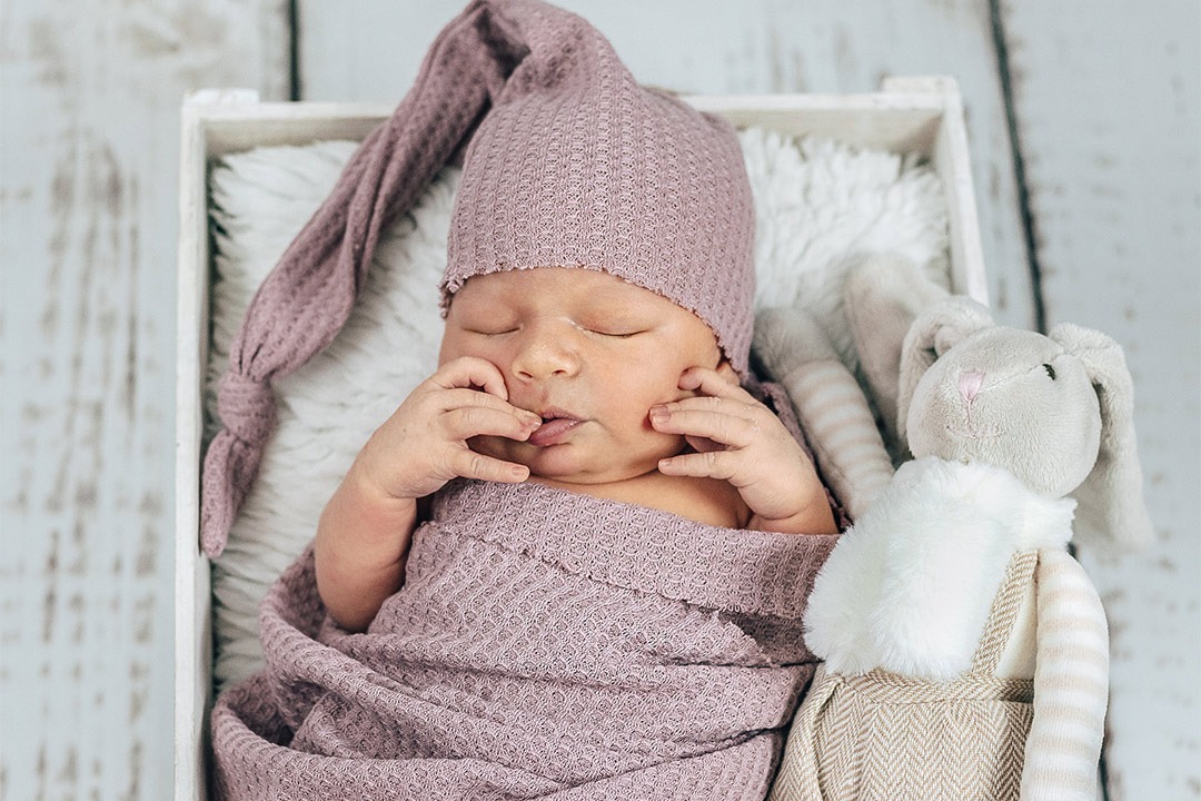 Śpiący noworodek w liliowym czepku i otulaczu, leżący w białej drewnianej skrzynce na miękkim futerku, obok pluszowy królik w beżowych ogrodniczkach.