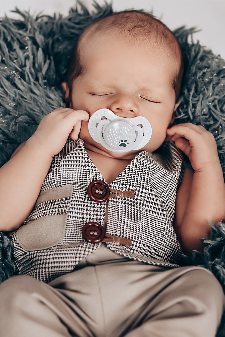 Śpiący noworodek w tweedowej kamizelce z drewnianymi guzikami i smoczkiem z nadrukiem łapy na tle szarego, futrzanego kocyka.