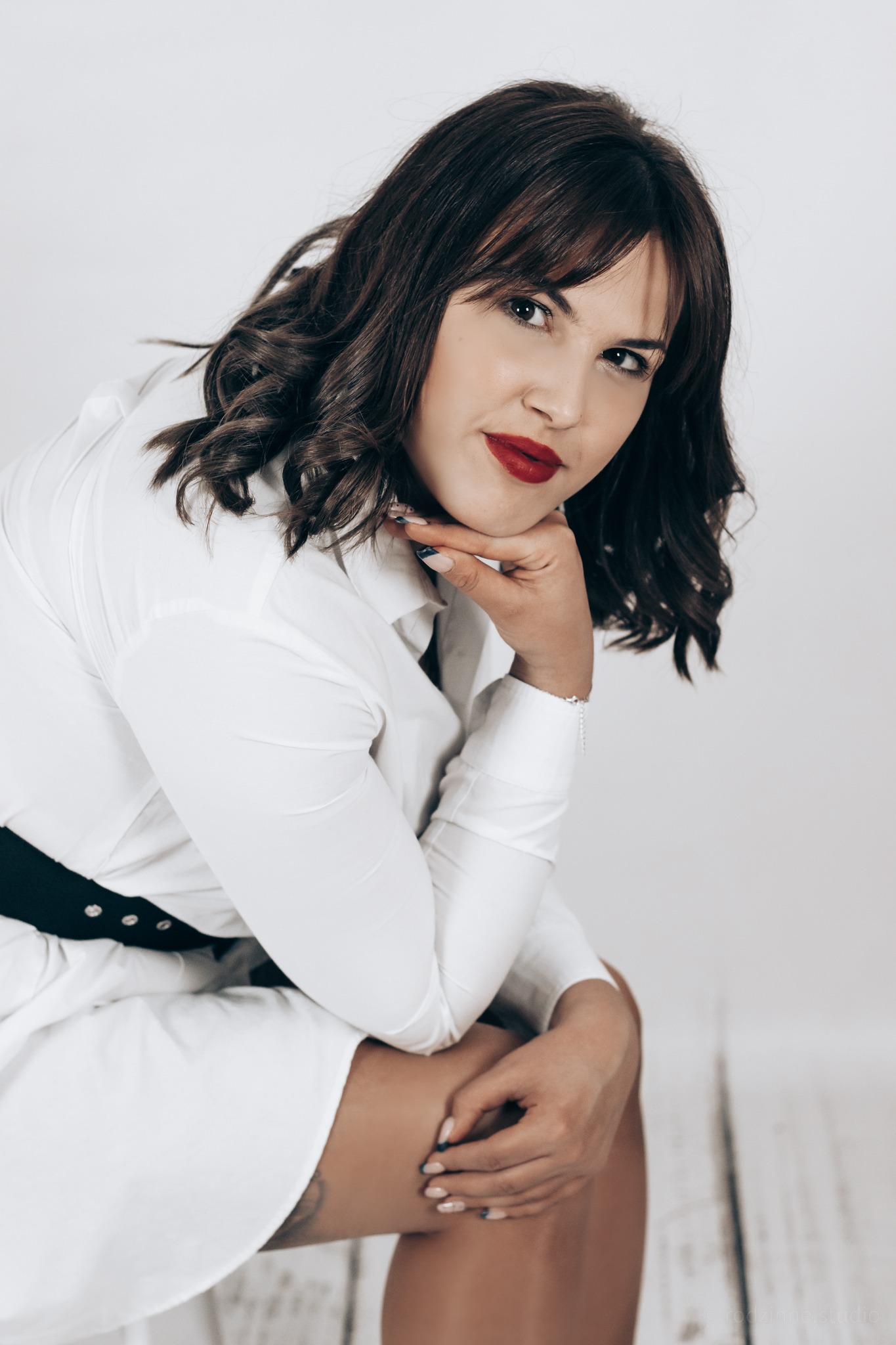 Kobieta w białej sukience koszulowej z czarnym paskiem, opierająca brodę na dłoni, z ciemnymi, kręconymi włosami i czerwoną szminką na ustach, pozująca na jasnym tle.