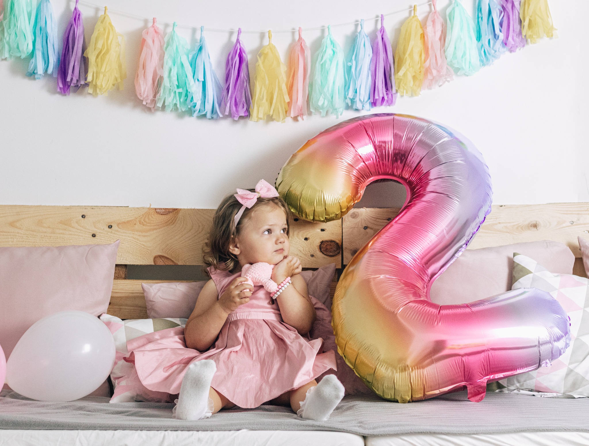 Dziewczynka w różowej sukience siedzi na łóżku z balonem w kształcie cyfry dwa i trzyma w ręku pluszaka; w tle girlanda z kolorowych frędzli.