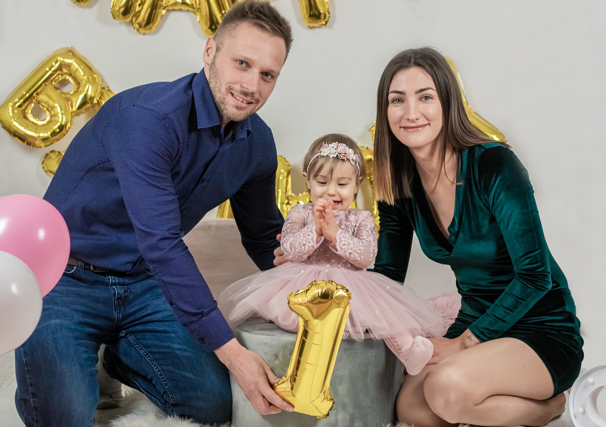Uśmiechnięta rodzina świętuje pierwsze urodziny dziecka: tata trzyma złoty balon w kształcie cyfry 1, mama i córka w różowej sukience siedzą na pufie, w tle biała ściana z balonami.