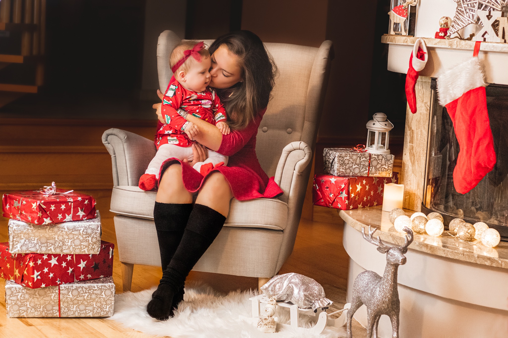 Kobieta w czerwonej sukience siedzi w fotelu i całuje w policzek małą dziewczynkę ubraną w świąteczny kombinezon, w tle kominek z dekoracjami i prezenty.