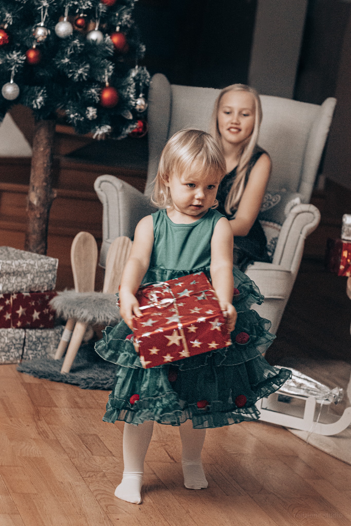 Dziewczynka w zielonej sukience niesie prezent w czerwonym papierze ze złotymi gwiazdami, w tle choinka i siedząca na fotelu starsza dziewczynka.