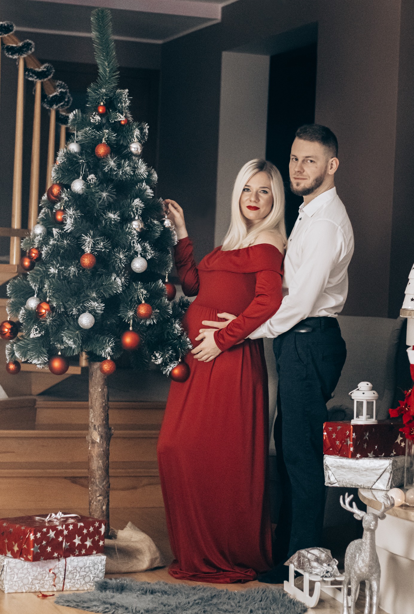 Ciężarna blondynka w czerwonej sukni off-shoulder z partnerem przy choince, świąteczna sesja w domowym klimacie.
