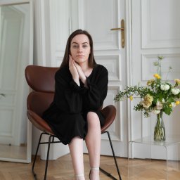 Dominik Stefanowski - Kobieta w czarnej sukience i jasnych butach siedzi na brązowym krześle, dłonie oparte o policzki, obok bukiet żółto-białych kwiatów na przezroczystym stoliku