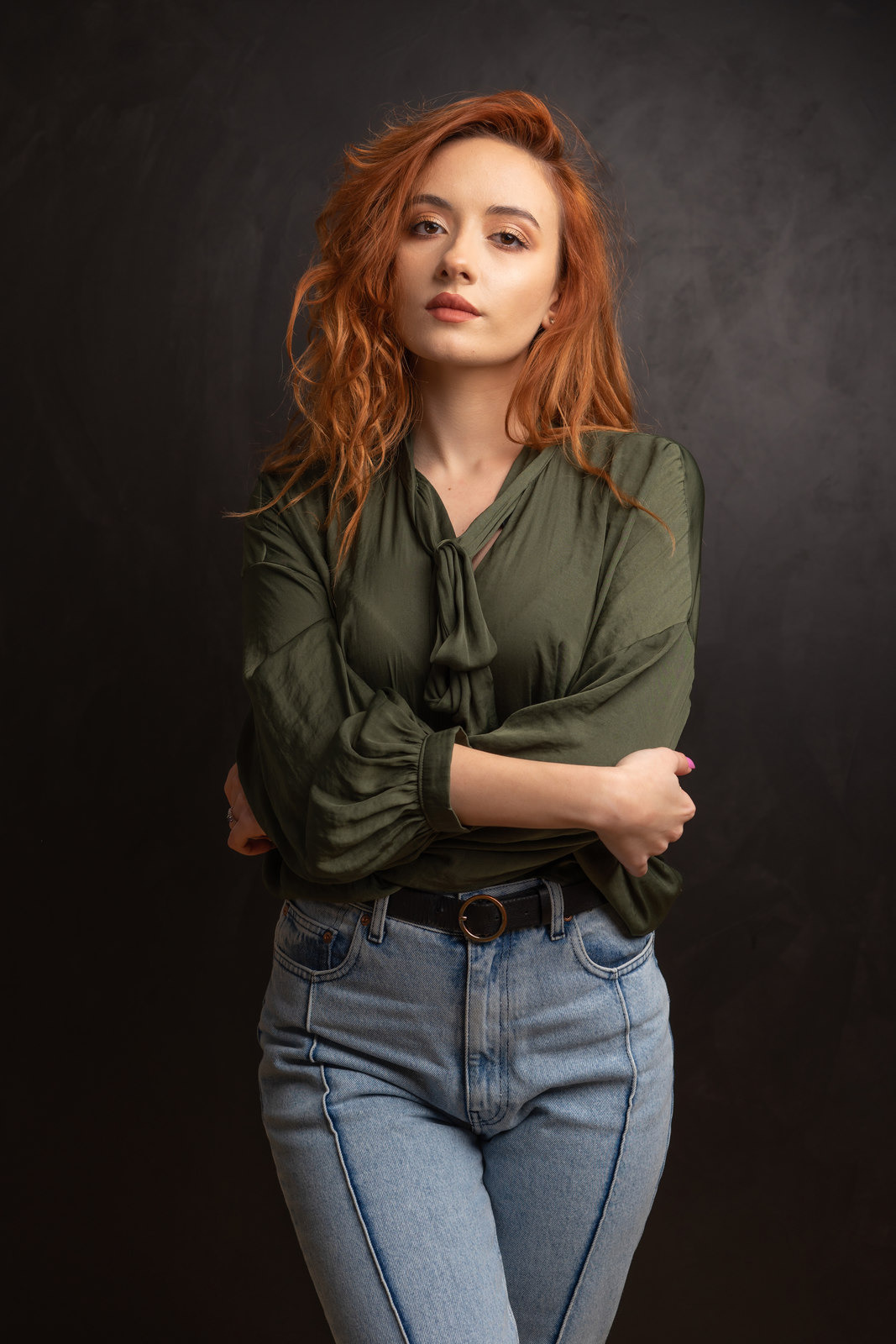 Portret kobiety z rudymi włosami, ubranej w oliwkową bluzkę i jeansy, pozującej na ciemnym tle z rękami skrzyżowanymi na piersiach.