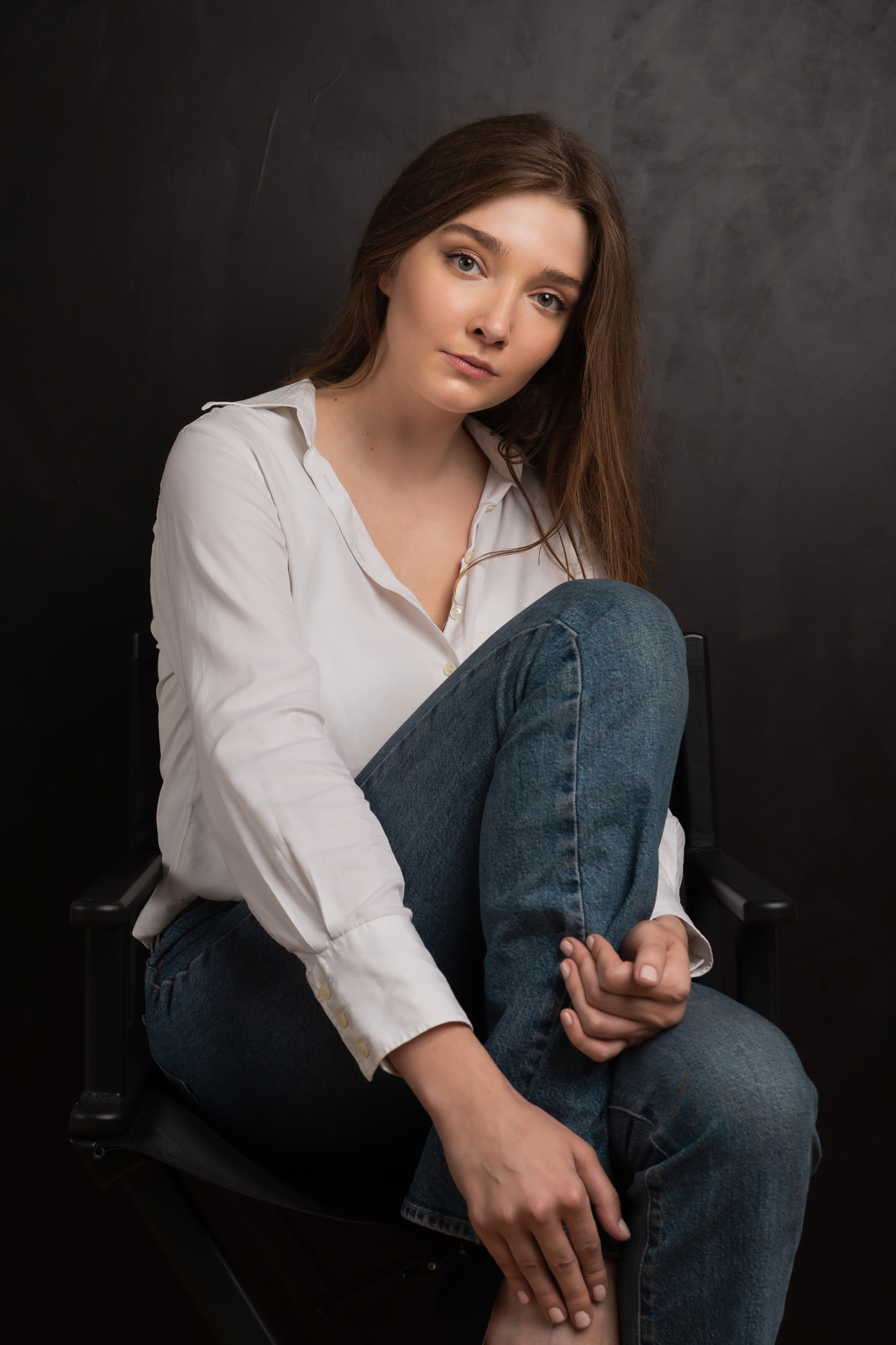Portret kobiety w białej koszuli i dżinsach siedzącej na krześle na ciemnym tle, z nogami podciągniętymi do góry i dłońmi splecionymi na kolanach, spoglądającej w obiektyw.