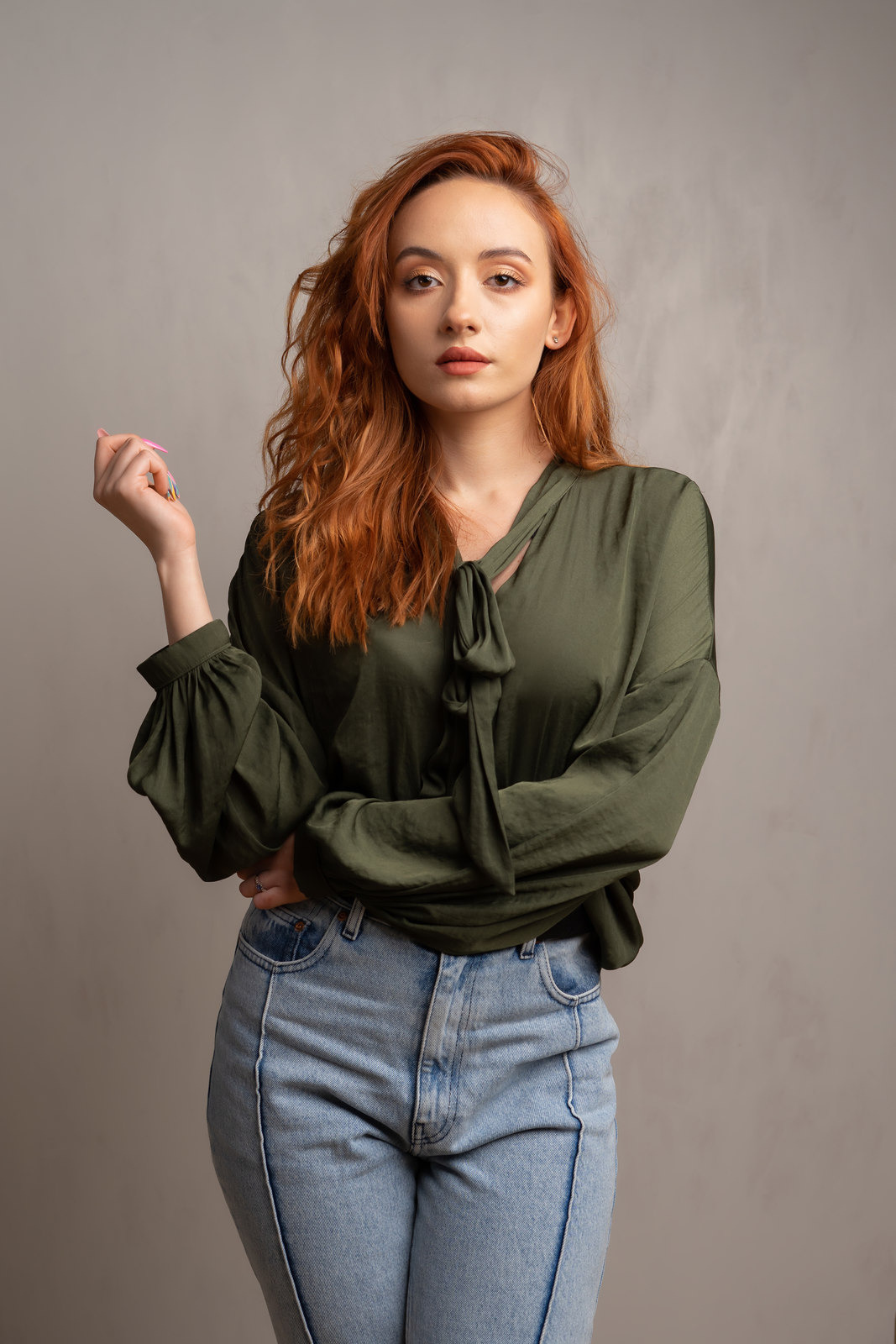 Portret kobiety z rudymi włosami w zielonej koszuli z kokardą i dżinsach, pozującej na szarym tle.