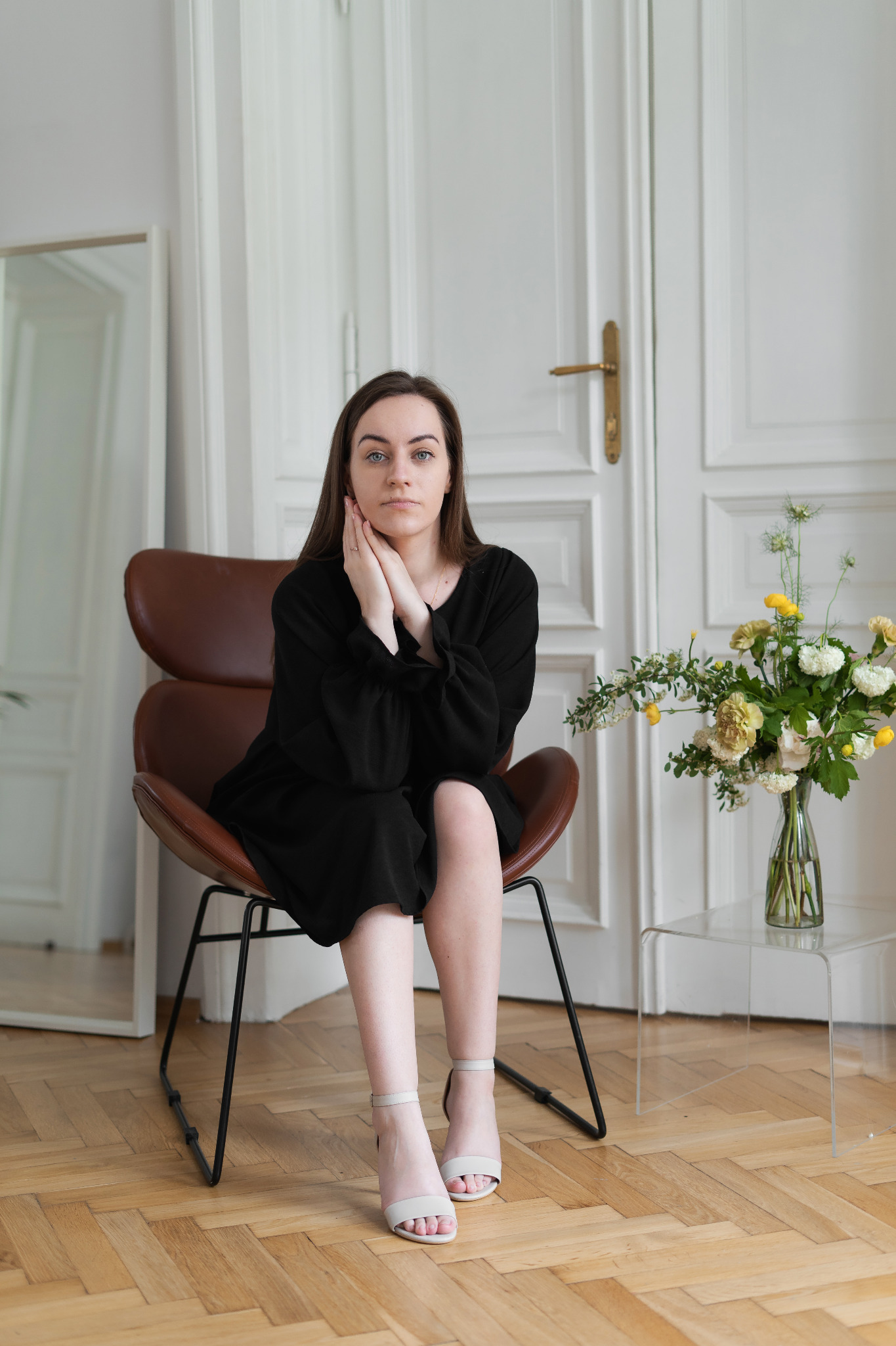 Kobieta w czarnej sukience i jasnych butach siedzi na brązowym krześle, dłonie oparte o policzki, obok bukiet żółto-białych kwiatów na przezroczystym stoliku