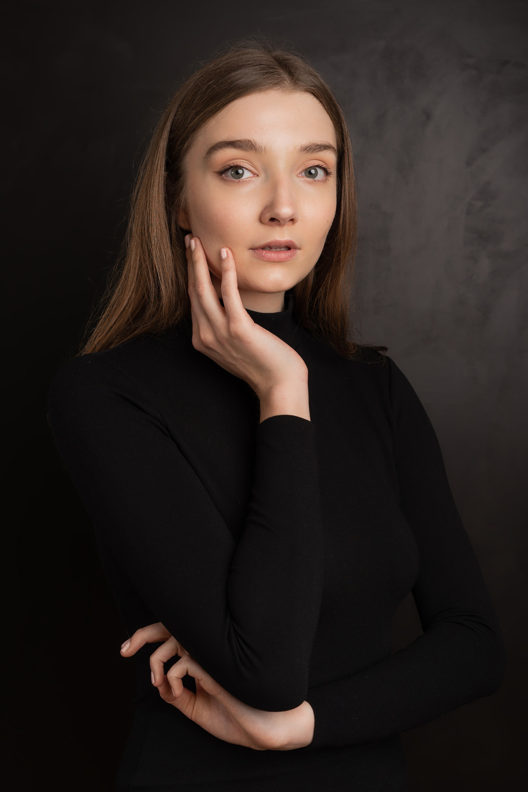 Portret kobiety w czarnym golfie, z ręką delikatnie dotykającą policzka, na ciemnym, rozmytym tle.