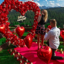 Balony Happy Day Zakopane - Zaręczyny w plenerze: mężczyzna klęczy z pierścionkiem przed zaskoczoną kobietą na tle serca z czerwonych róż, balonów w kształcie serc, świec i górskiego krajobrazu, dekoracja z napisem neonowym...