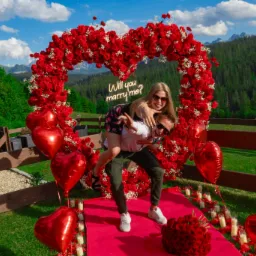 Zaręczyny w plenerze: mężczyzna trzyma na barana kobietę przed sercem z czerwonych róż z neonowym napisem 'Will you marry me?', czerwony dywan z płatkami róż i świecami, w tle górski krajobraz.