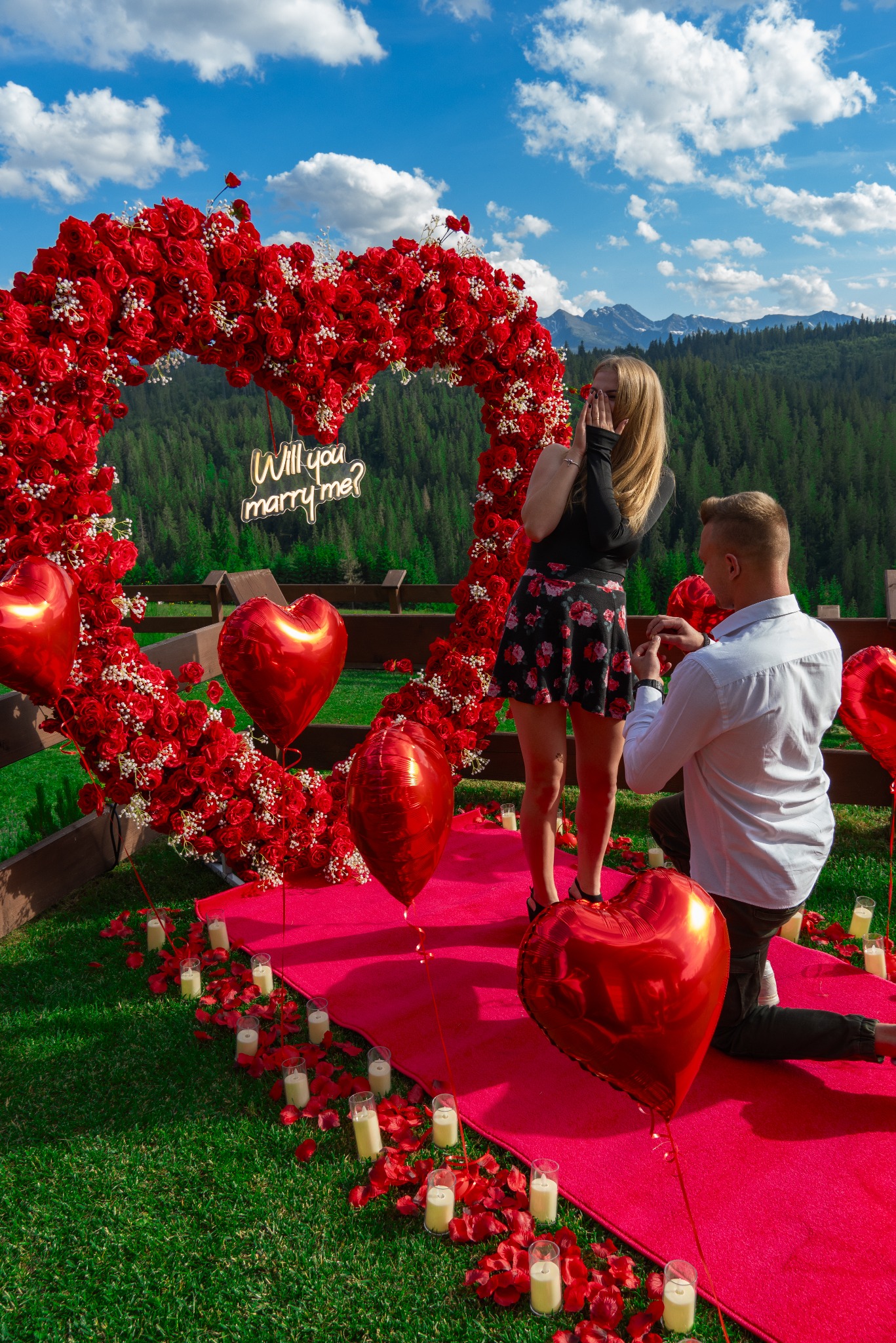 Zaręczyny w plenerze: mężczyzna klęczy z pierścionkiem przed zaskoczoną kobietą na tle serca z czerwonych róż, balonów w kształcie serc, świec i górskiego krajobrazu, dekoracja z napisem neonowym...