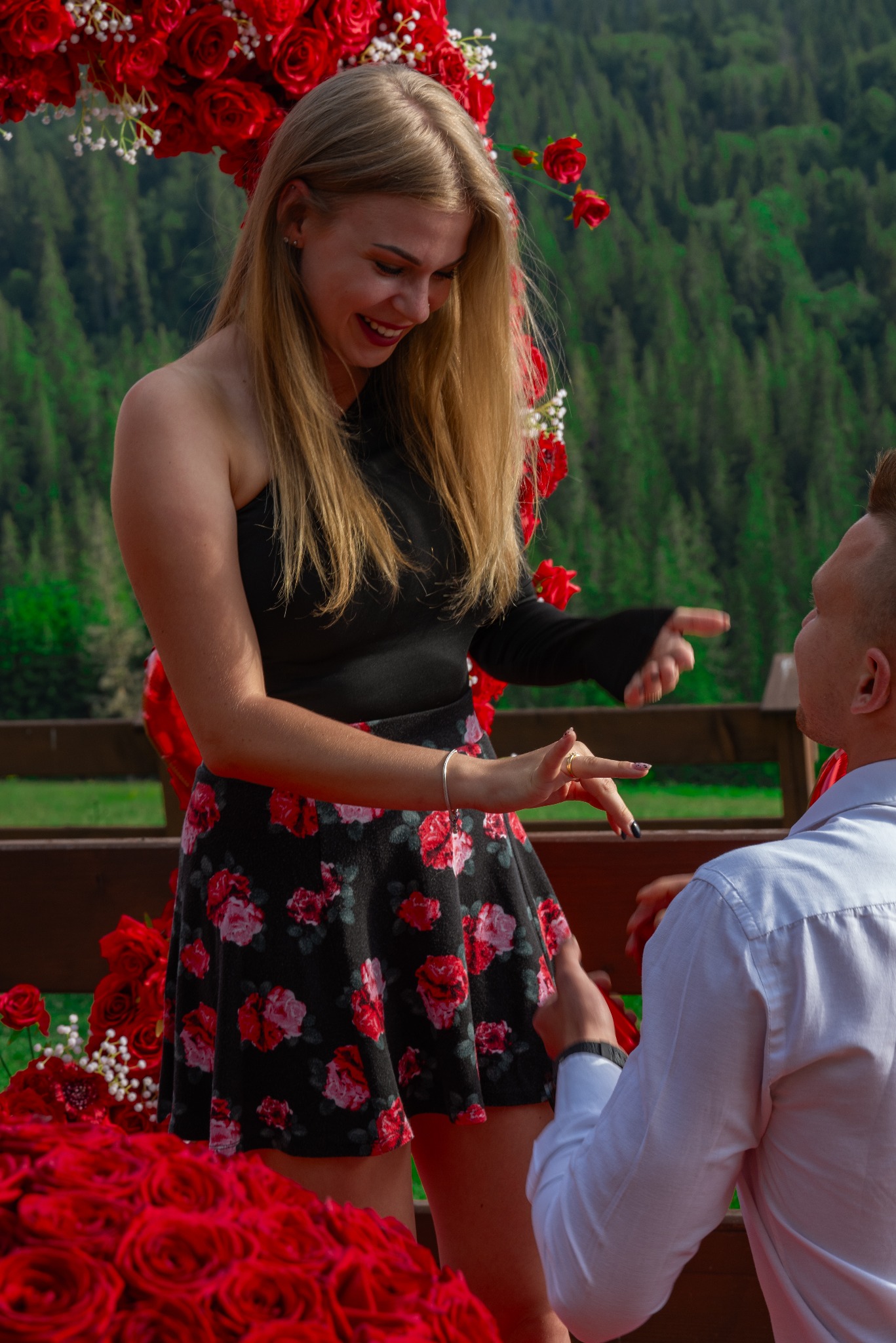 Zaręczyny w plenerze: uśmiechnięta kobieta z blond włosami i pierścionkiem na palcu, przyjmuje oświadczyny od mężczyzny na tle dekoracji z czerwonych róż i zalesionych wzgórz.