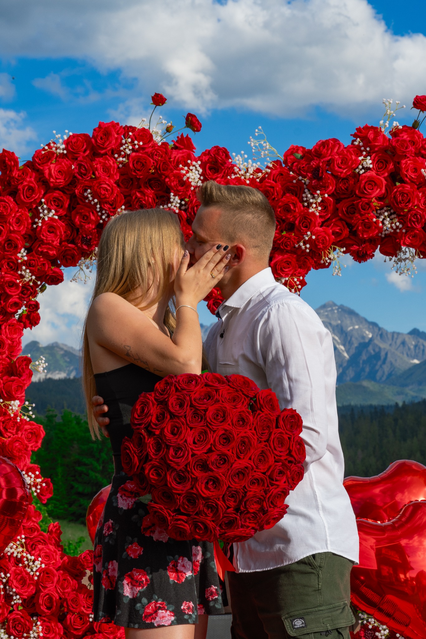 Para całująca się na tle gór i łuku z czerwonych róż, mężczyzna trzyma bukiet z czerwonych róż, zaręczyny w plenerze.