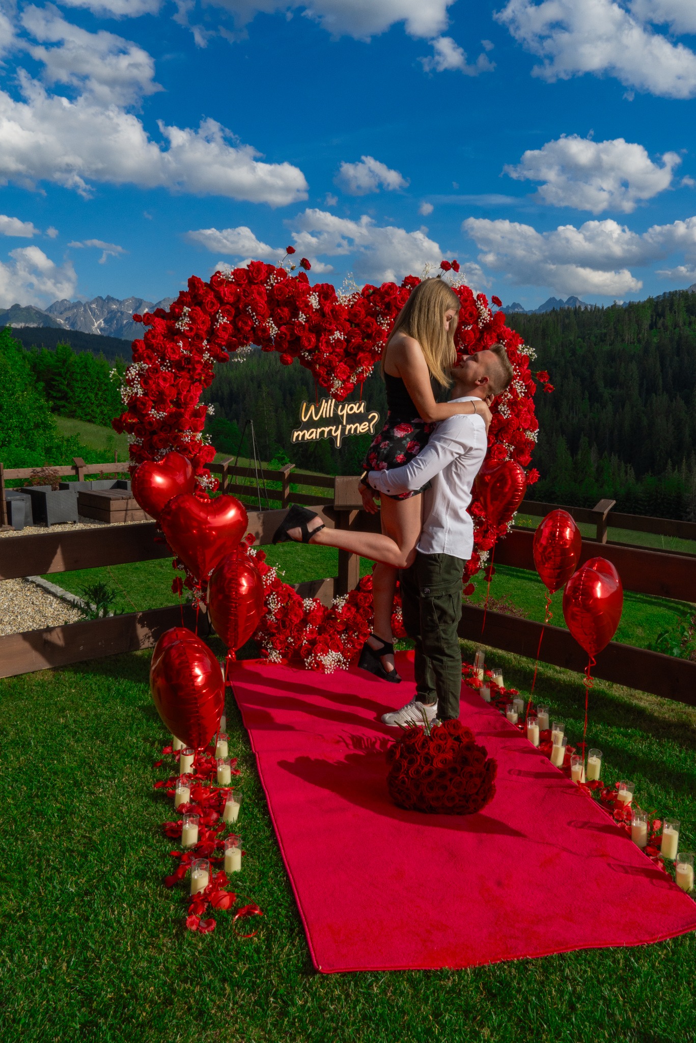 Mężczyzna trzyma na rękach kobietę przed sercem z czerwonych róż z napisem neonowym 'Will you marry me?', czerwony dywan, świece i płatki róż na trawie, w tle górski krajobraz.