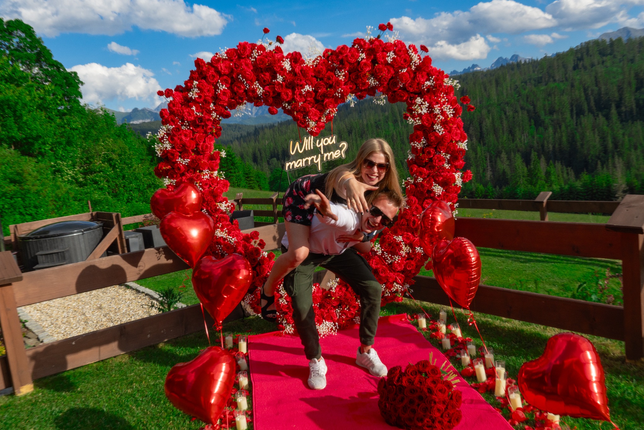 Zaręczyny w plenerze: mężczyzna trzyma na barana kobietę przed sercem z czerwonych róż z neonowym napisem 'Will you marry me?', czerwony dywan z płatkami róż i świecami, w tle górski krajobraz.