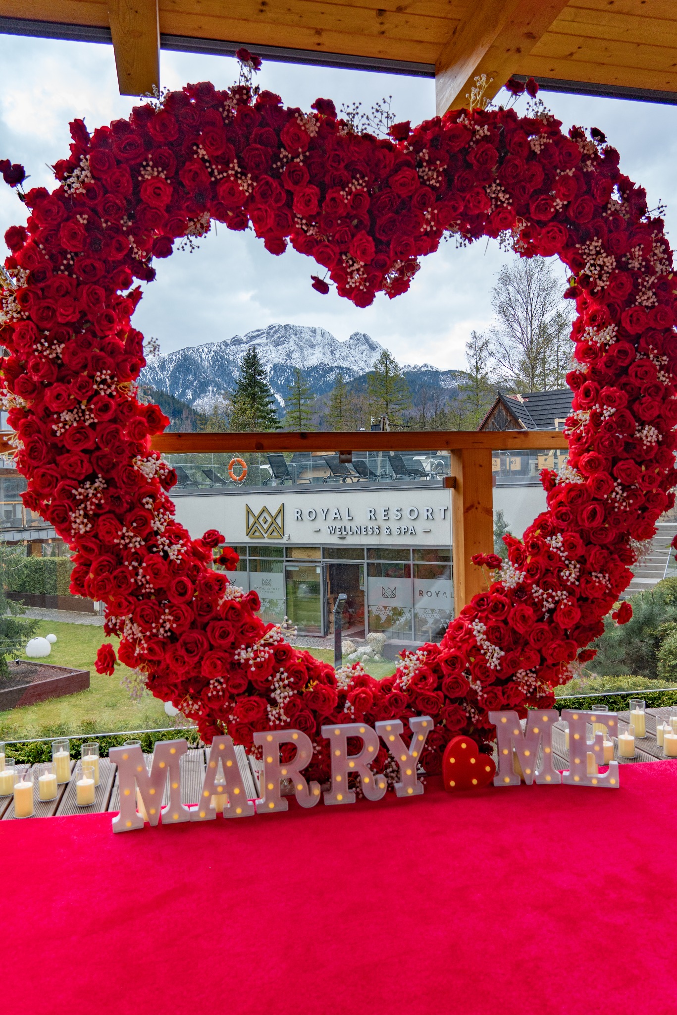 Romantyczna sceneria zaręczyn w Zakopanem: serce z czerwonych róż i białych gipsówek, podświetlany napis 'MARRY ME', świece, czerwony dywan, w tle budynek Royal Resort Wellness & Spa oraz ośnieżone...