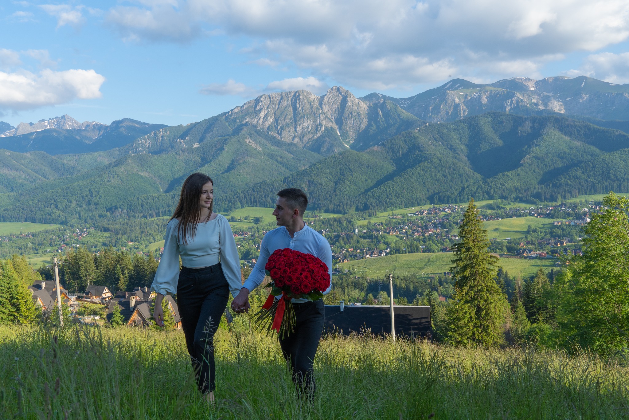 Para trzymająca się za ręce spaceruje po łące z bukietem czerwonych róż na tle górskiego krajobrazu Zakopanego.