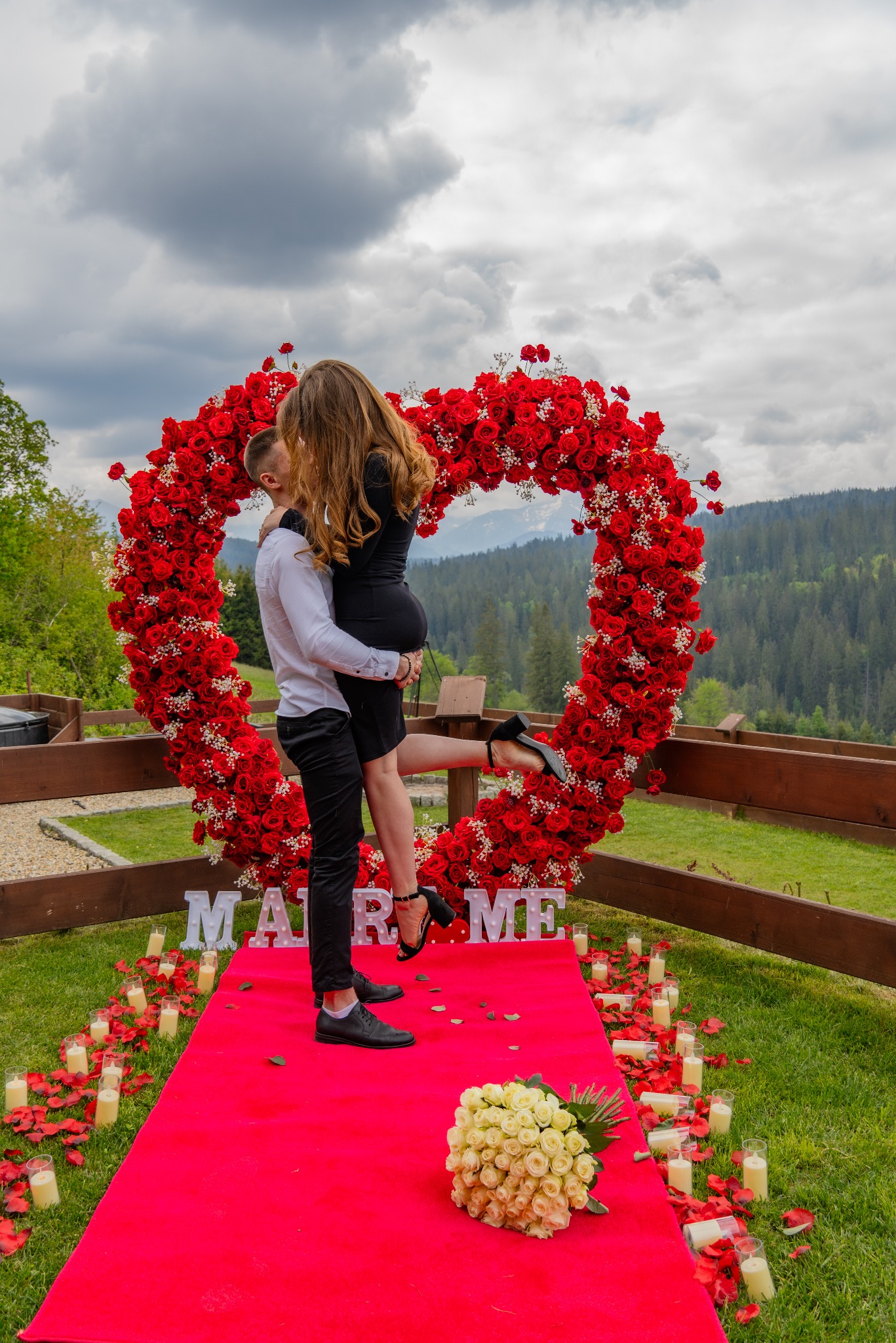 Para całująca się na tle serca z czerwonych róż, z napisem 'MARRY ME' i świecami, na tle górskiego krajobrazu.