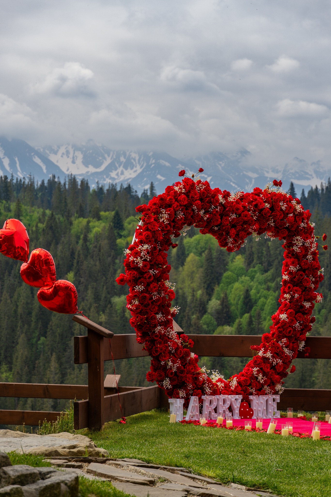 Romantyczna sceneria zaręczyn na tle gór: serce z czerwonych róż, balony w kształcie serc, podświetlane litery 'MARRY ME' i świece na zielonej trawie.