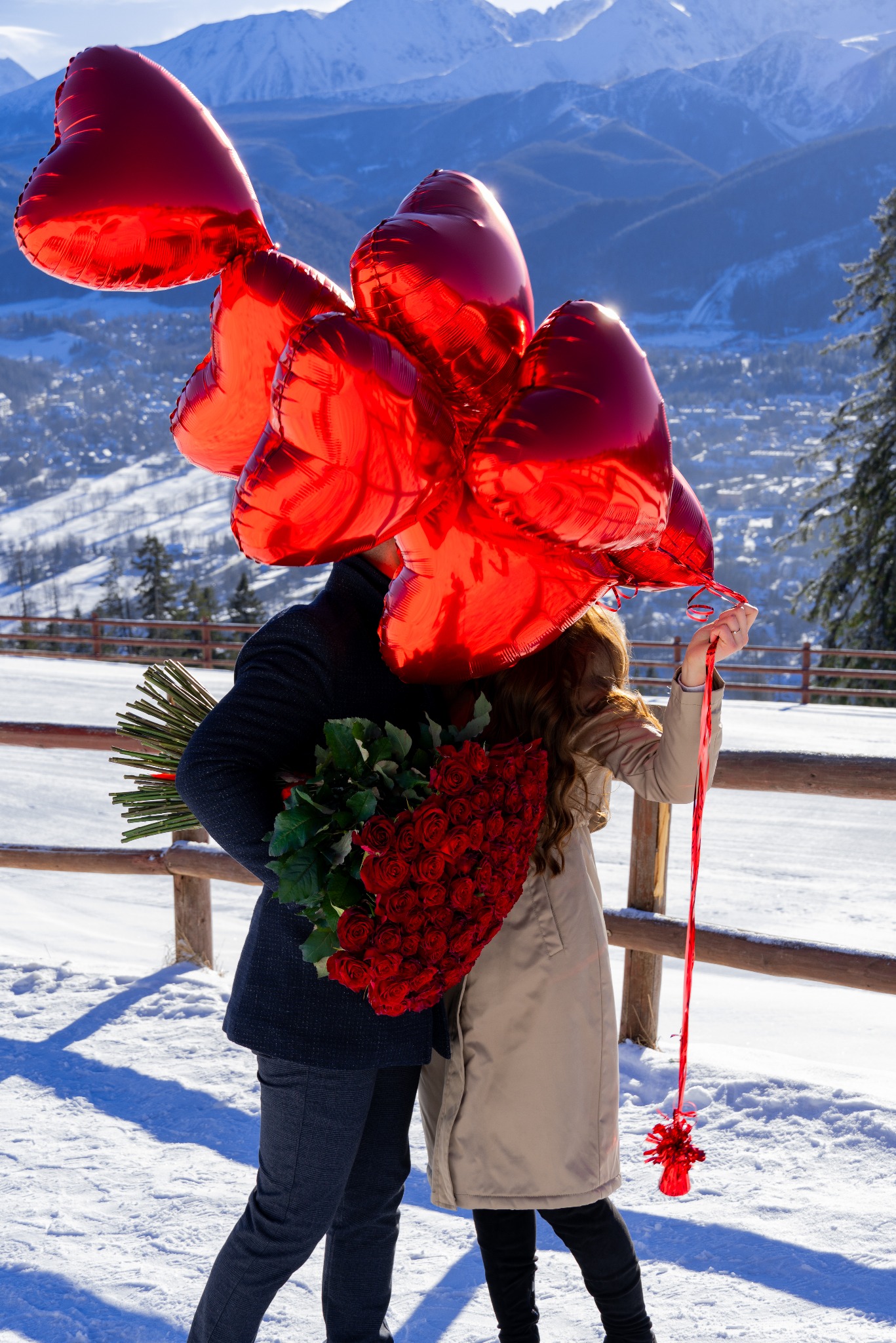 Para na tle górskich szczytów zasłonięta bukietem czerwonych róż i czerwonymi balonami w kształcie serc.