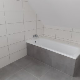 ES-BUD Eryk Szczerbowski - Wykończona łazienka na poddaszu z białą wanną obudowaną szarymi płytkami, białe płytki na ścianach i szara podłoga, widoczne gniazdko elektryczne i bateria wannowa.