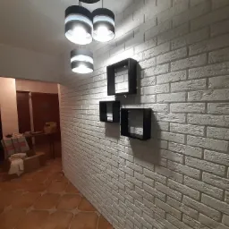 Ściana z białej cegły dekoracyjnej z trzema czarnymi, kwadratowymi półkami i nowoczesną lampą sufitową