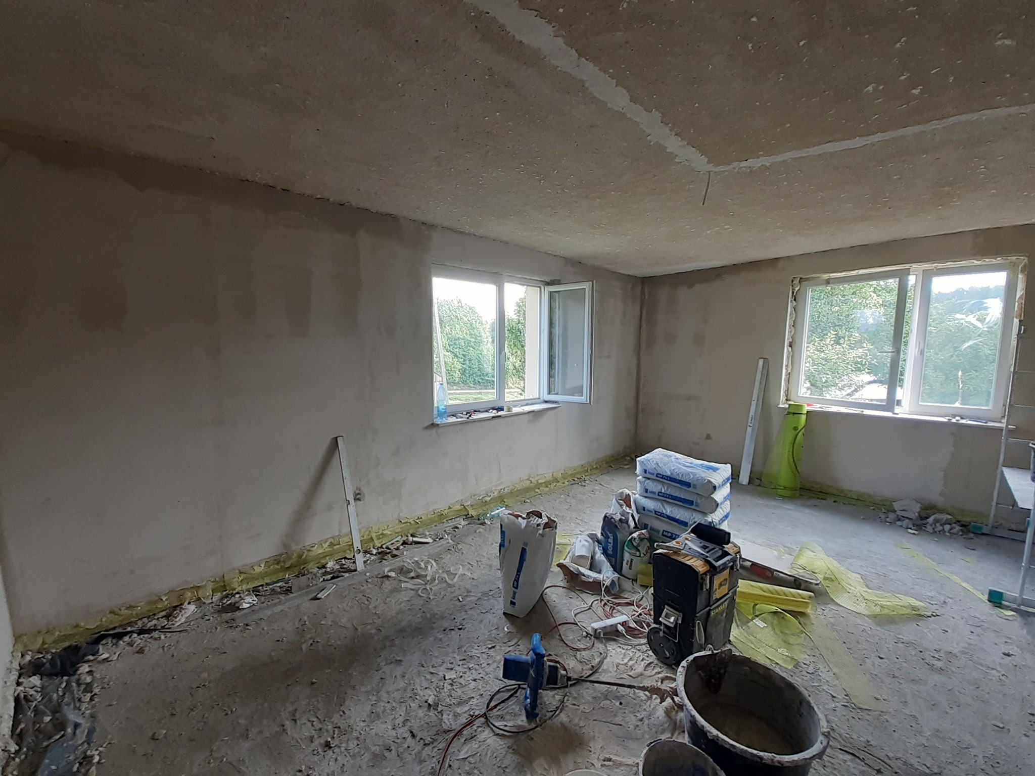 Pokój w trakcie prac wykończeniowych: surowe ściany i sufit, worki z zaprawą, narzędzia budowlane i gruz na podłodze, widok z okna na zieleń.