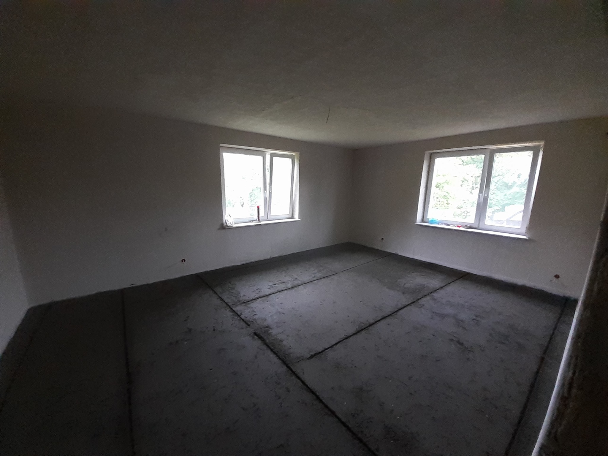 Pokój w stanie surowym: wylewka podłogowa z widocznymi dylatacjami, surowe ściany i sufit, dwa okna z widokiem na zieleń.