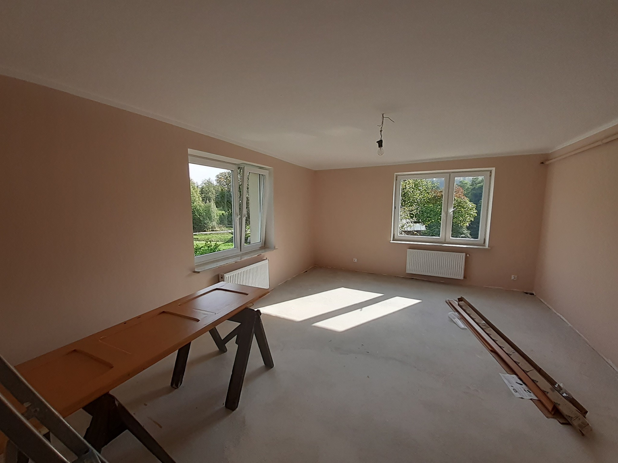 Pokój po malowaniu ścian na kolor brzoskwiniowy, z drewnianymi listwami przypodłogowymi w opakowaniu, drzwiami opartymi na kozłach malarskich i widokiem na zieleń za oknami.