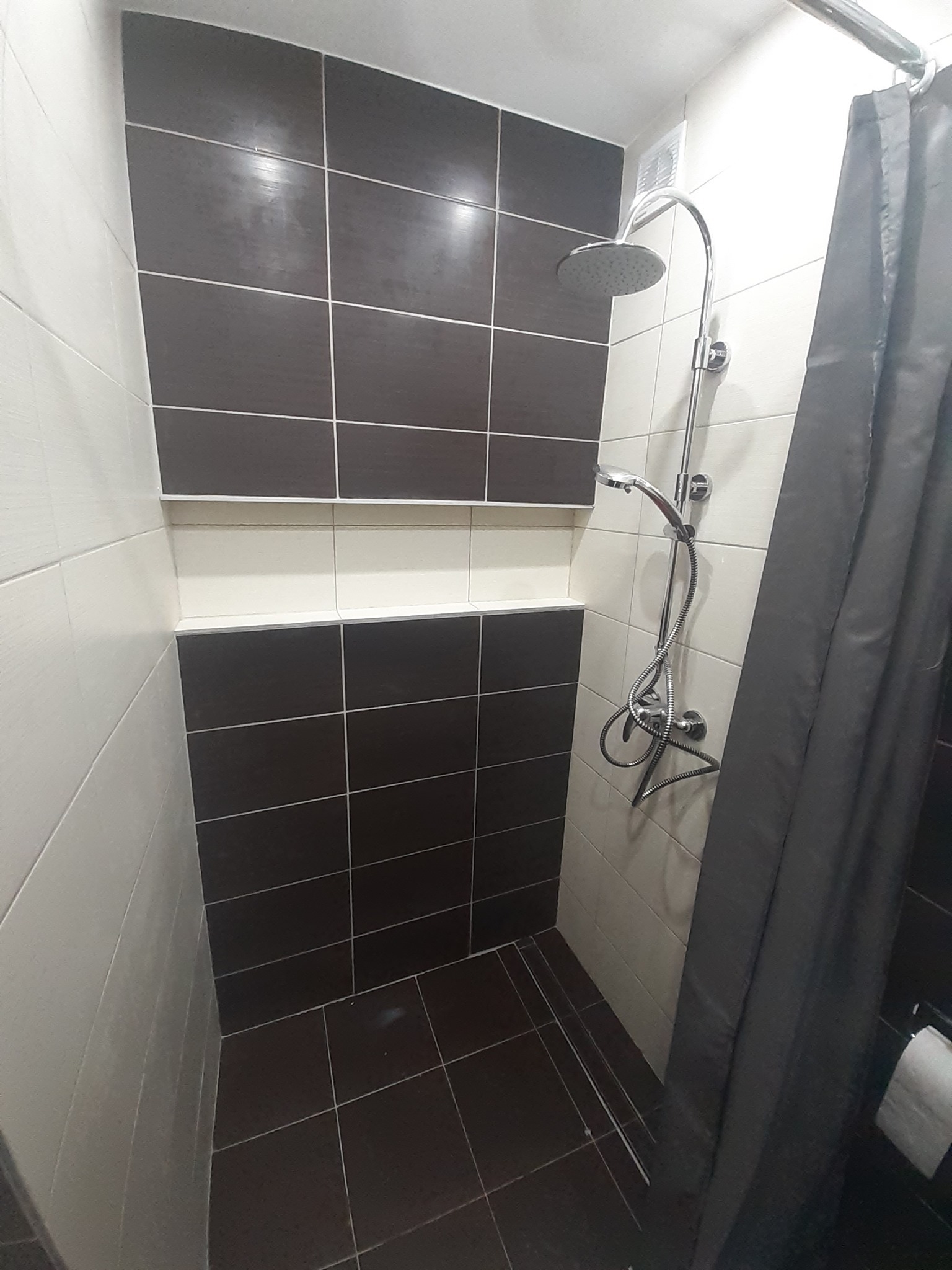 Kabina prysznicowa z chromowaną armaturą, wykończona płytkami w odcieniach brązu i beżu, z szarą zasłoną prysznicową.