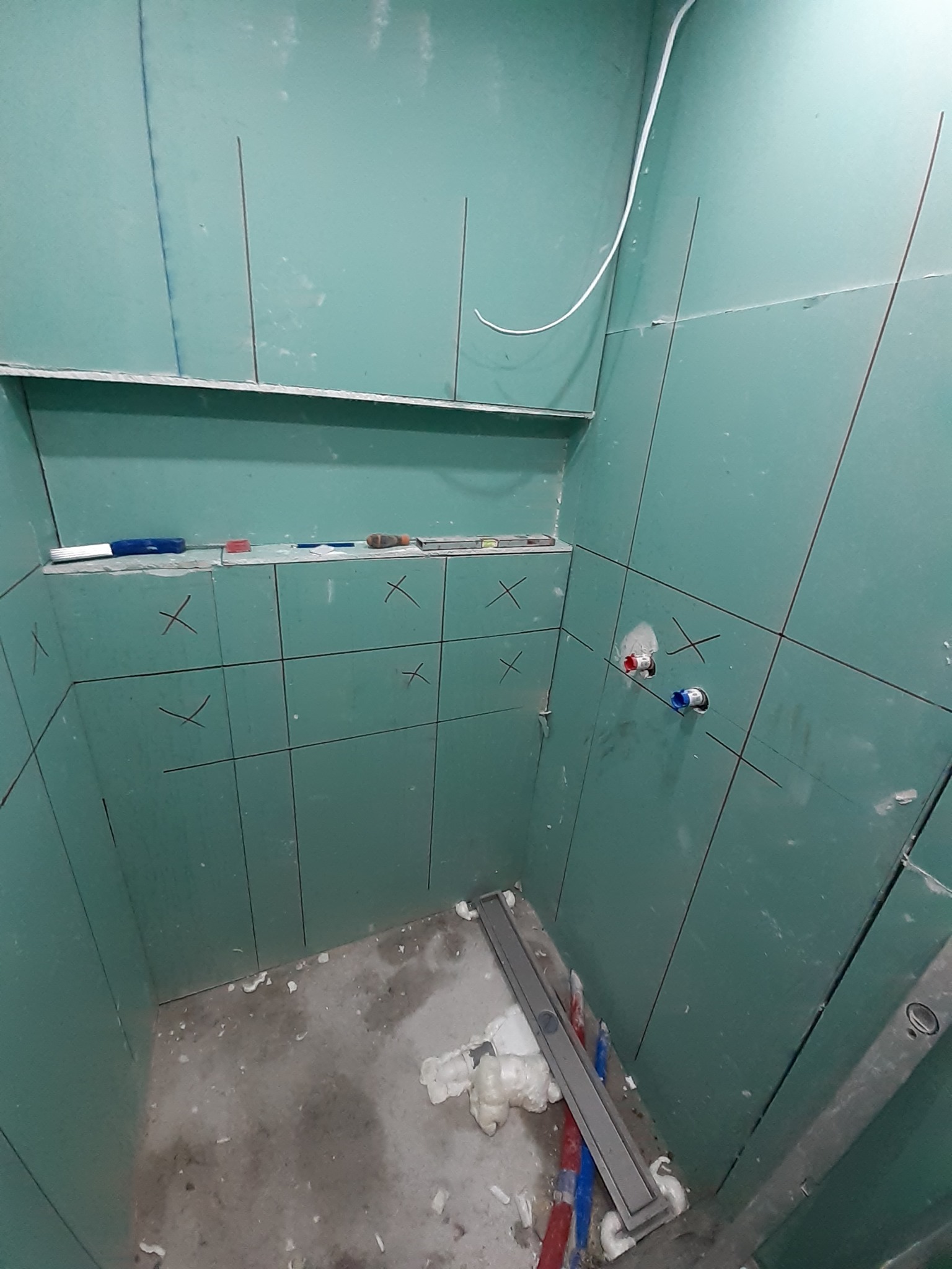 Przygotowanie ścian w łazience do położenia płytek: zielone płyty gipsowo-kartonowe z zaznaczonymi liniami cięcia, widoczne elementy instalacji hydraulicznej i odpływ liniowy w podłodze, narzędzia...