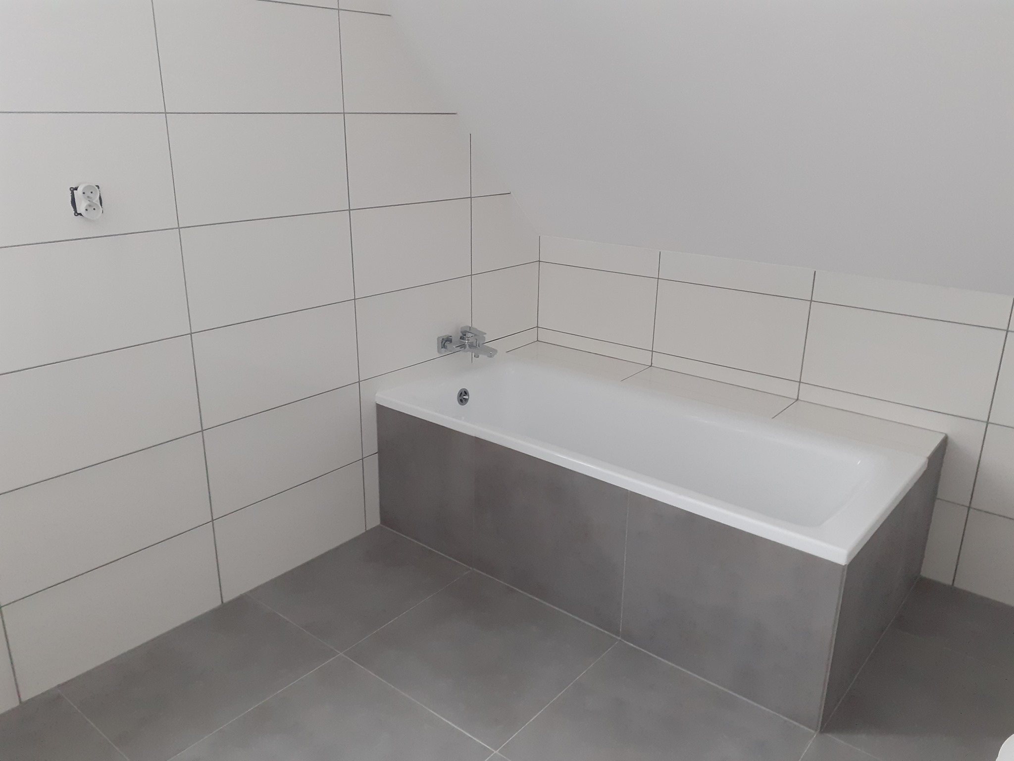 Wykończona łazienka na poddaszu z białą wanną obudowaną szarymi płytkami, białe płytki na ścianach i szara podłoga, widoczne gniazdko elektryczne i bateria wannowa.