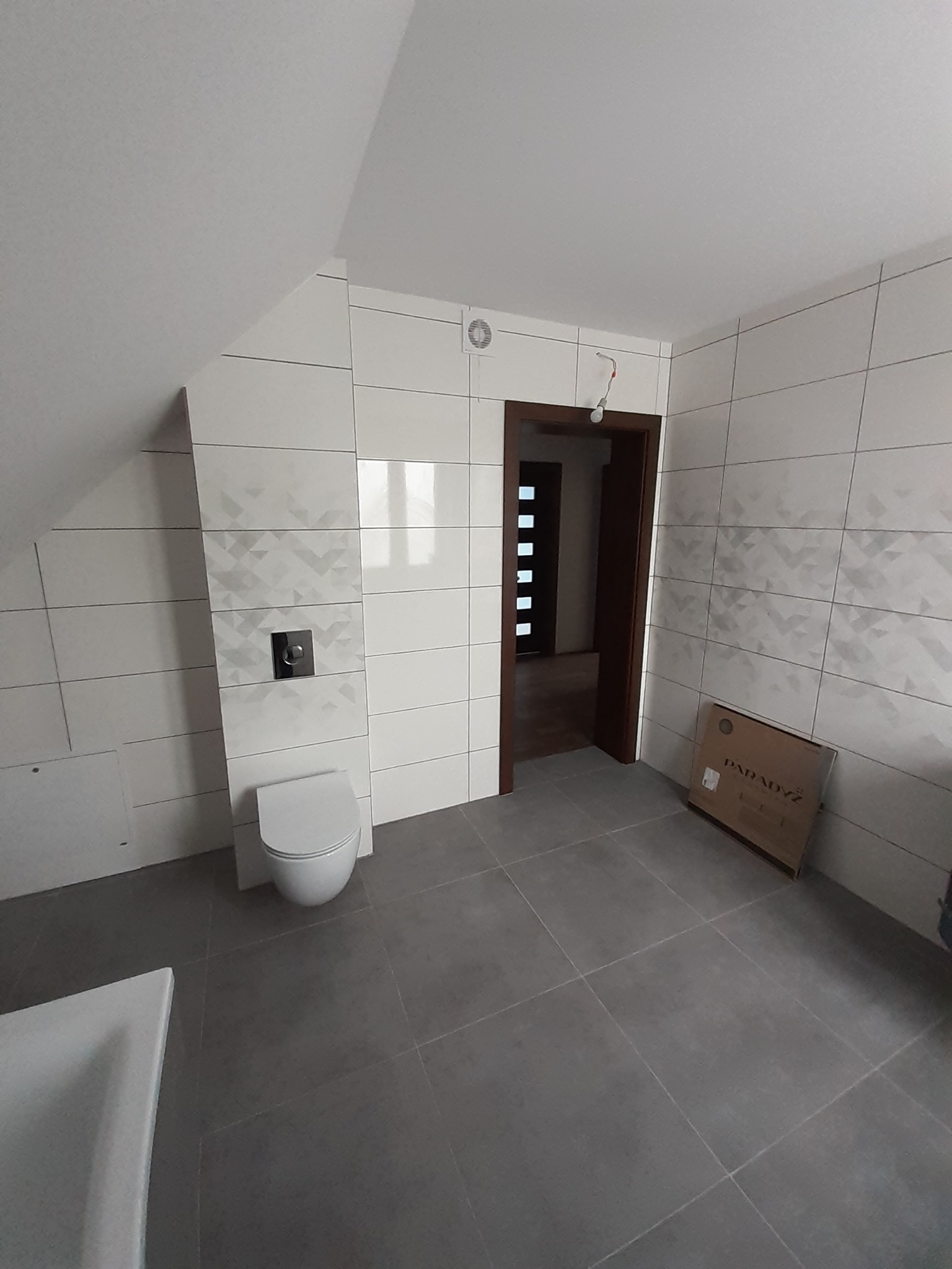 Wykończona łazienka na poddaszu z białymi płytkami ściennymi z delikatnym wzorem geometrycznym, szarą podłogą, podwieszaną toaletą i otwartymi drzwiami do innego pomieszczenia, widoczne kartony po...