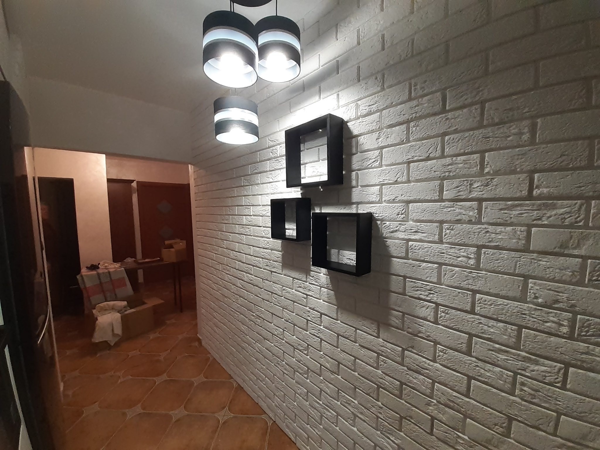 Ściana z białej cegły dekoracyjnej z trzema czarnymi, kwadratowymi półkami i nowoczesną lampą sufitową