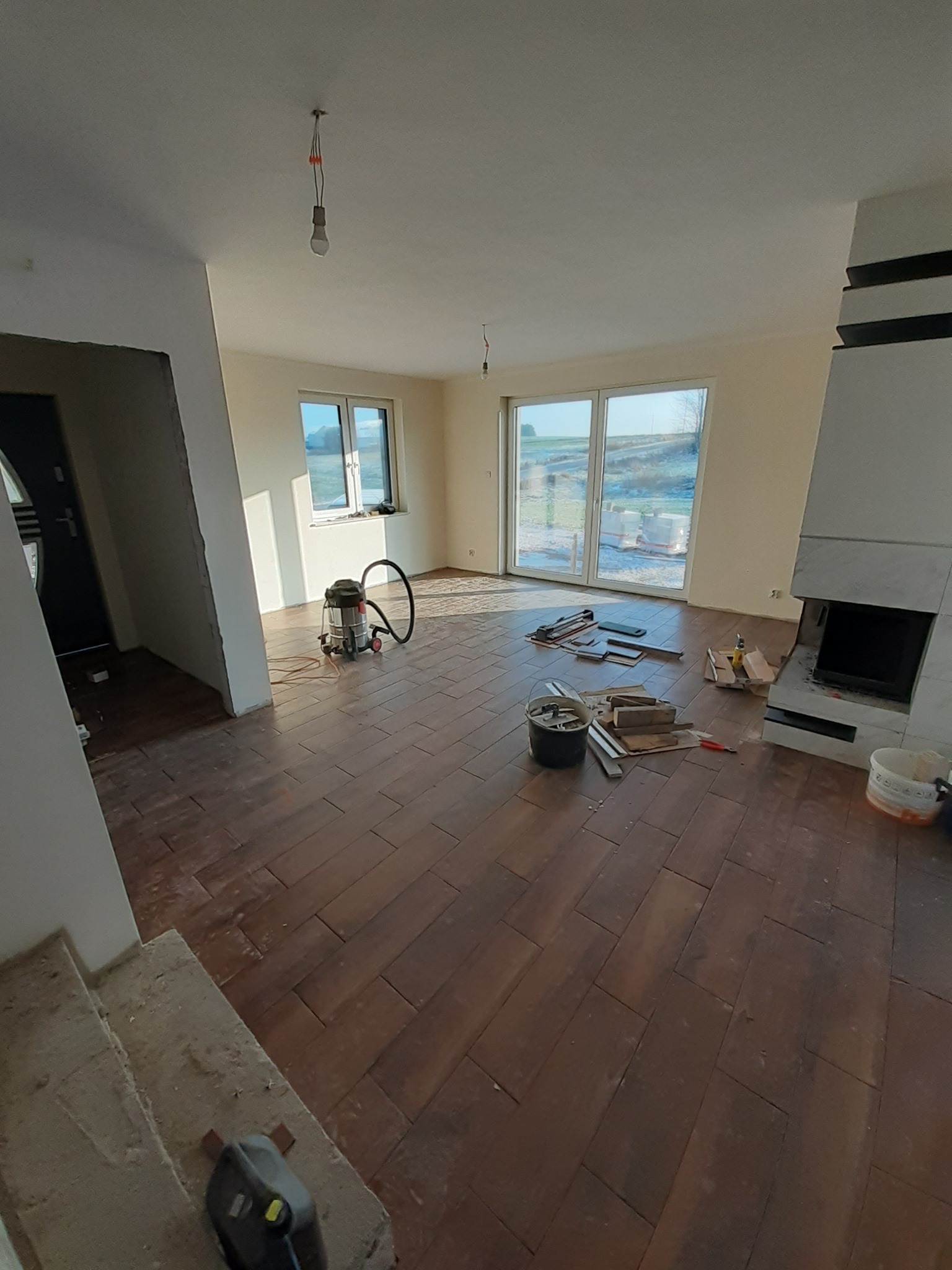 Wnętrze w trakcie układania ciemnobrązowej podłogi z płytek imitujących drewno, widoczne narzędzia, odkurzacz przemysłowy, kominek i okno z widokiem na zewnątrz.