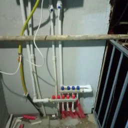 Rozdzielacz ogrzewania podłogowego z rurami w kolorach czerwonym i białym, zamontowany na balkonie, widoczne fragmenty rur i narzędzi na podłodze, z boku balustrada balkonu.