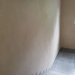 Wnętrze domu w trakcie budowy, widoczne otynkowane ściany w odcieniach beżu, szare schody wyłożone płytkami i fragment surowego sufitu.