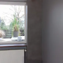 Wnętrze pokoju z oknem: ściana pomalowana na szaro z efektem dekoracyjnym w rogu, biały grzejnik pod oknem, za oknem widok na balkon i drzewa.