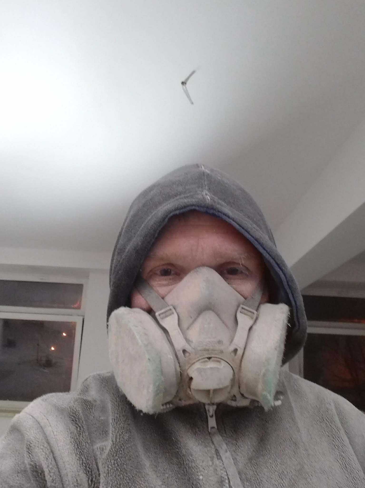 Mężczyzna w masce przeciwpyłowej i bluzie z kapturem, pokryty pyłem, patrzący w obiektyw; widoczny fragment sufitu z zamontowanym wiatrakiem.